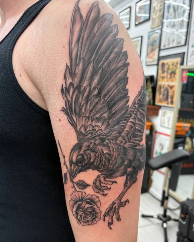 Bird and rose tattoo by Heather Baldwin — sparrow clutching a rose in its beak, neo-traditional black and grey on the upper arm