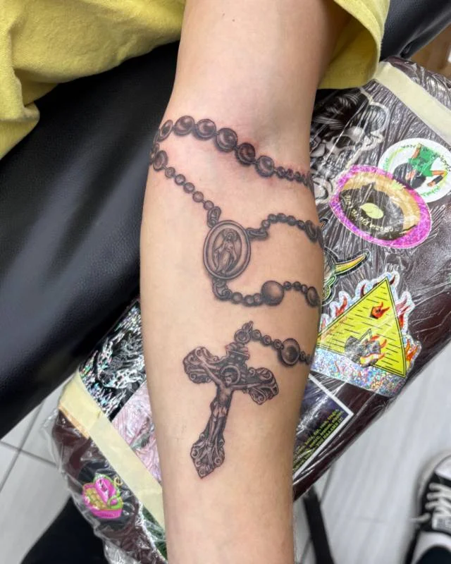Rosary and religious tattoo by Heather Baldwin — wrapped rosary with crucifix and Mary medallion on the forearm, black and grey realism
